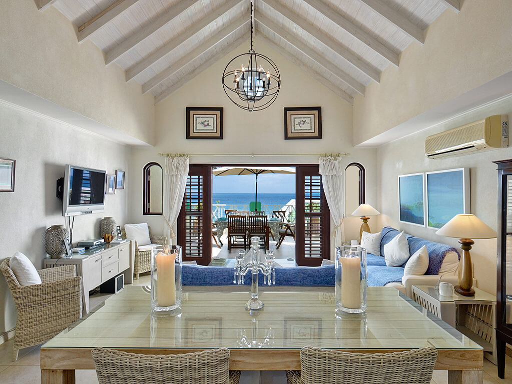 A luxurious living room with high vaulted ceilings, featuring a dining table with candles, wicker chairs, a blue sofa, and a TV. Large windows and glass doors open to a balcony with ocean views, framed by white curtains. Elegant decor and artwork adorn the space.