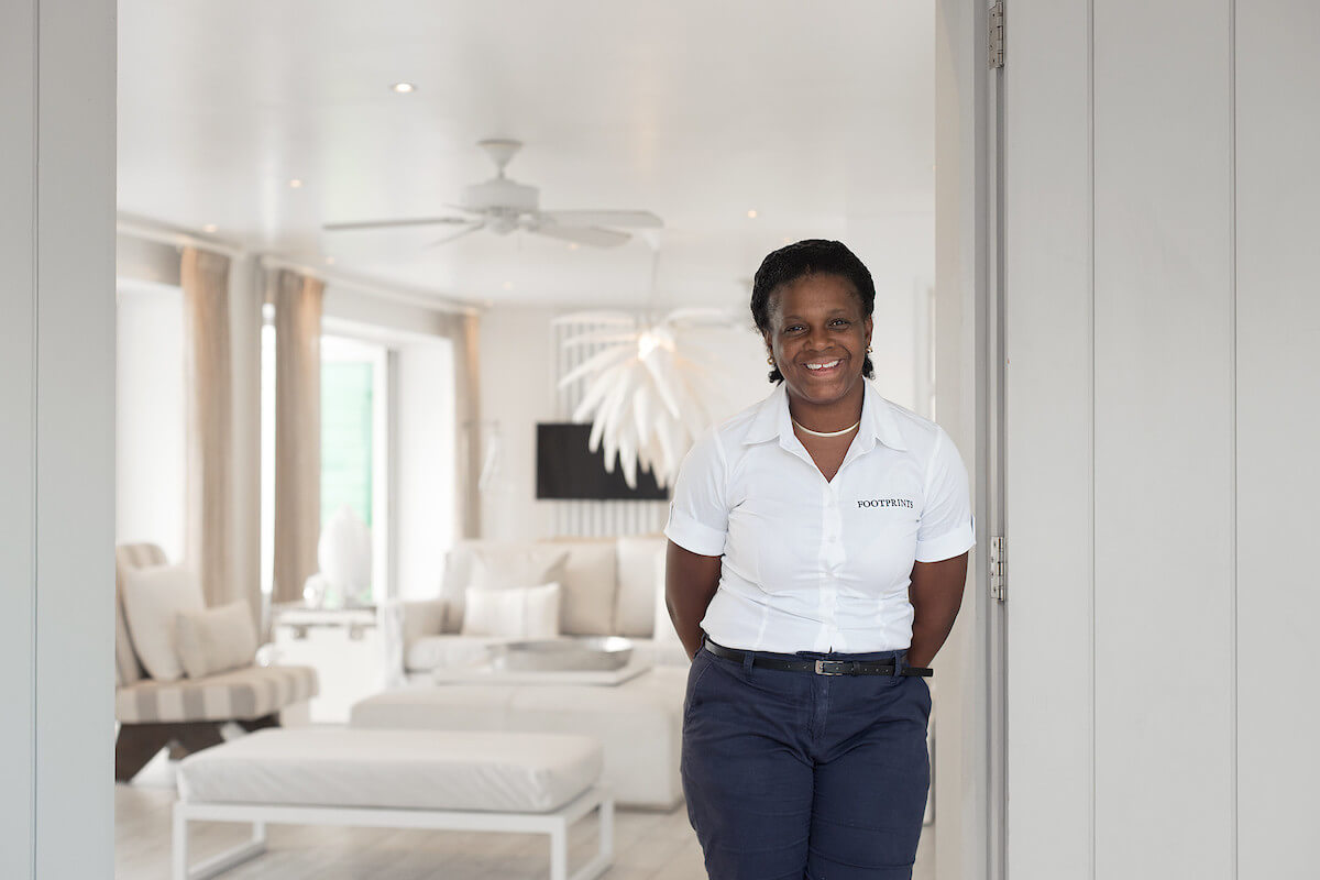 A smiling woman in a white uniform shirt and dark pants stands in the doorway of a bright, modern living room with white furniture and decor in a luxury Barbados holiday estate.