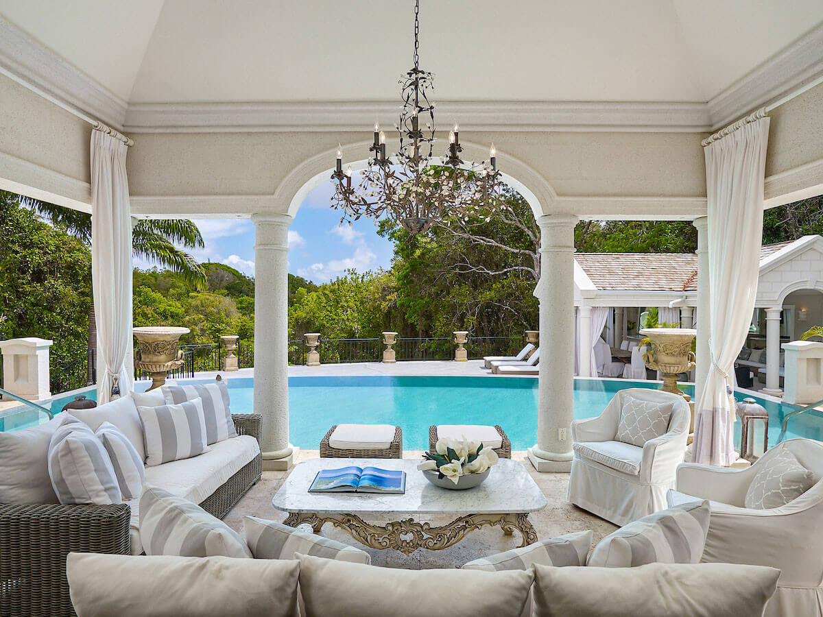 Luxurious covered patio with white sofas and armchairs overlooks an elegant infinity pool surrounded by greenery. Chandeliers hang from the ceiling, and an open book and flower arrangement rest on a marble coffee table.