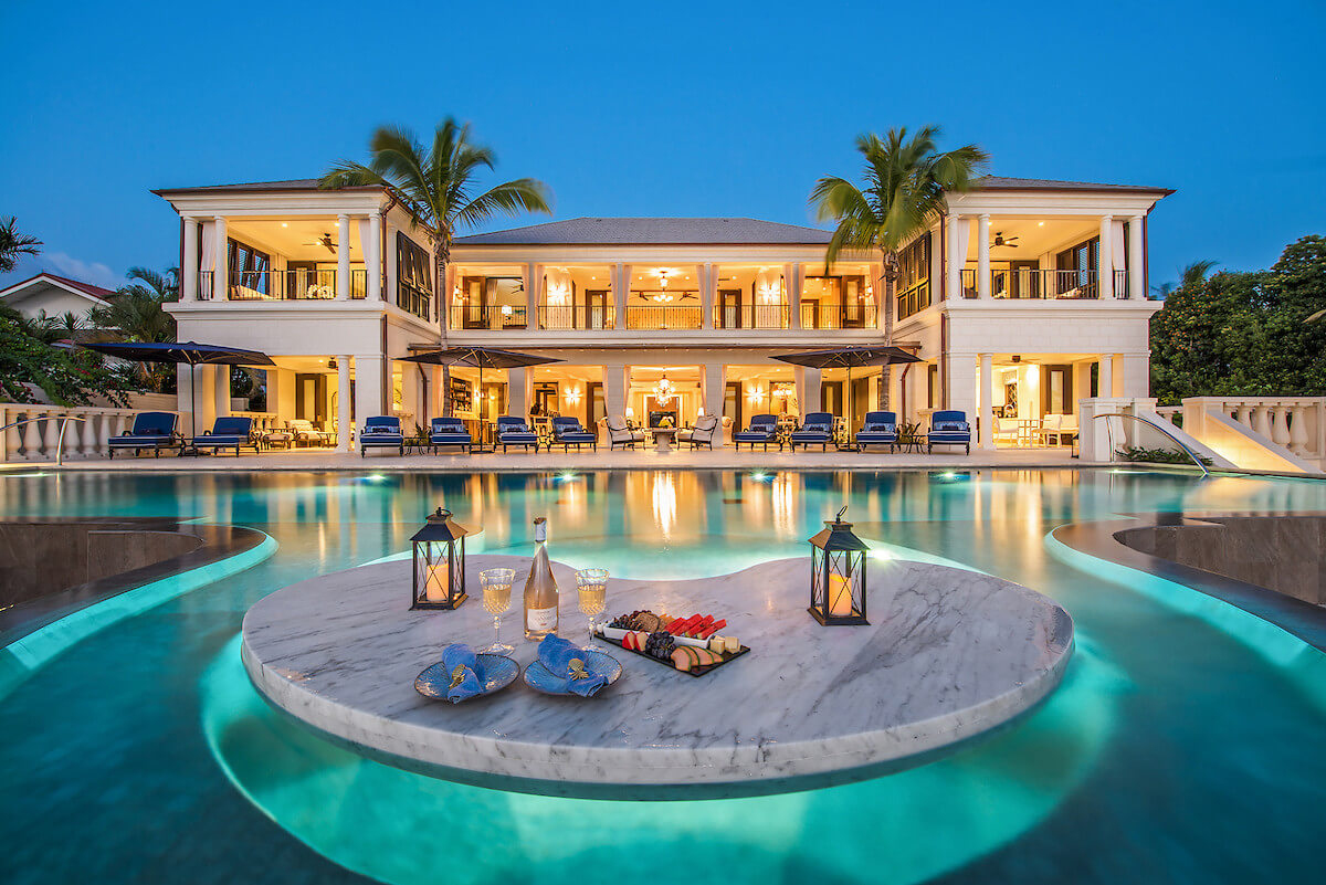 This six bedroom beachfront luxury villa in Barbados features balconies and large windows warmly lit at dusk, overlooking an expansive pool. A round marble table with snacks, drinks, and lanterns floats in the water, framed by swaying palm trees.