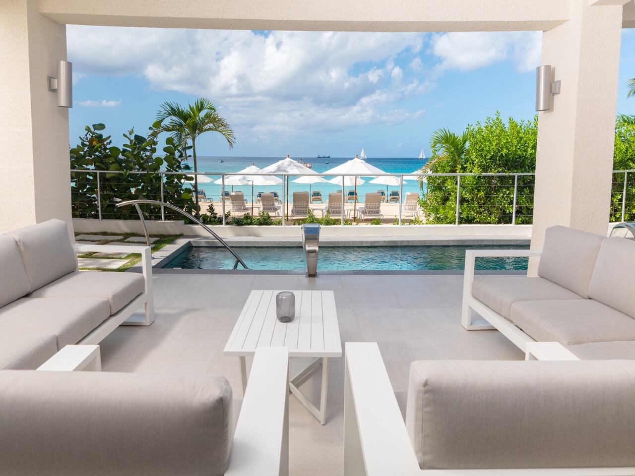A modern patio with light gray sofas and a white table overlooks a private pool, umbrellas, lounge chairs, and a beach with turquoise water under a partly cloudy sky.