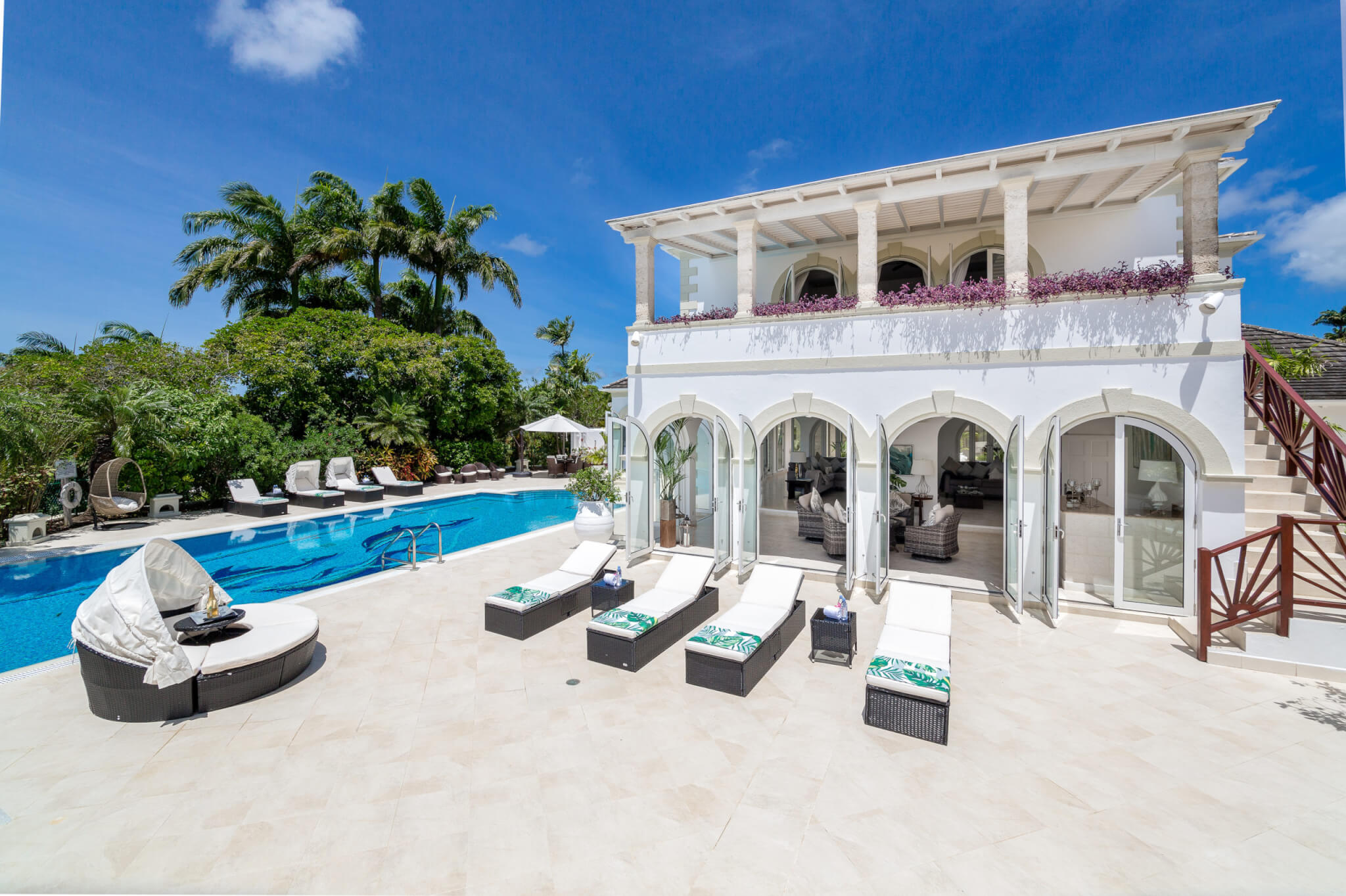 A luxurious white villa with a balcony overlooks a large swimming pool, surrounded by palm trees, lounge chairs, and sunny blue skies. Floor-to-ceiling windows open onto a spacious patio area.
