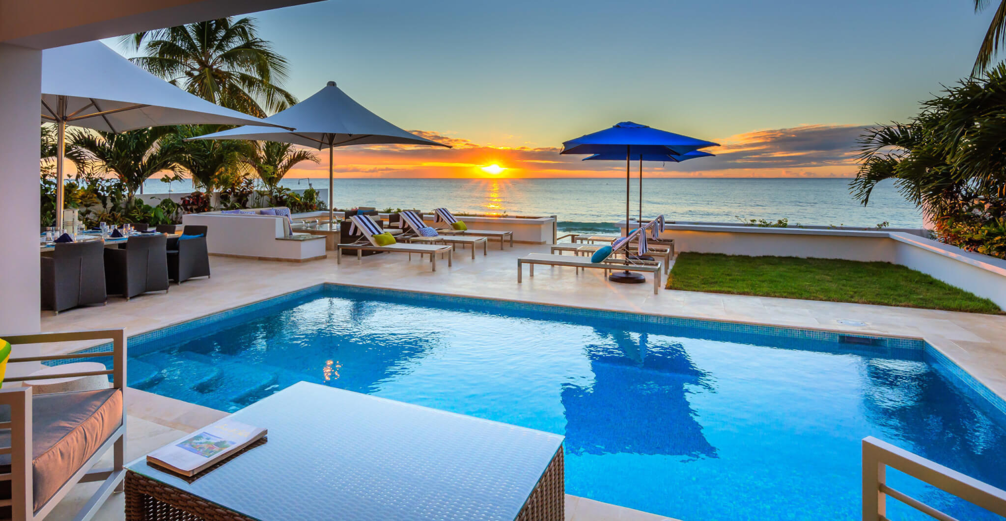 Luxury beachfront patio with a pool, lounge chairs, umbrellas, and palm trees overlooking the ocean at sunset. The sky is colorful, and the area is set up for relaxation and outdoor dining.