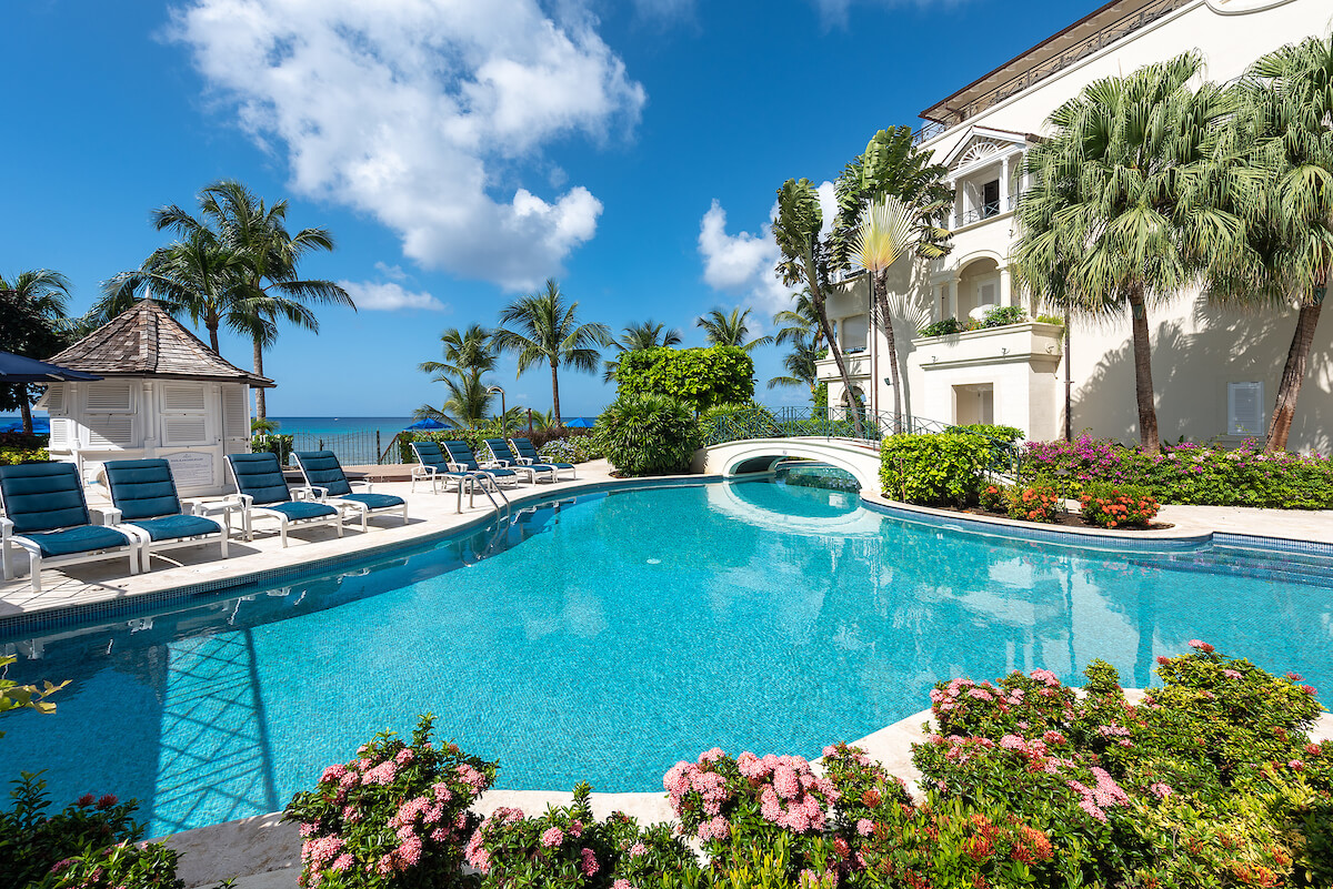 Luxury resort pool surrounded by lounge chairs, palm trees, and tropical flowers, with a white building and a clear blue sky in the background, and the ocean visible beyond the pool area.
