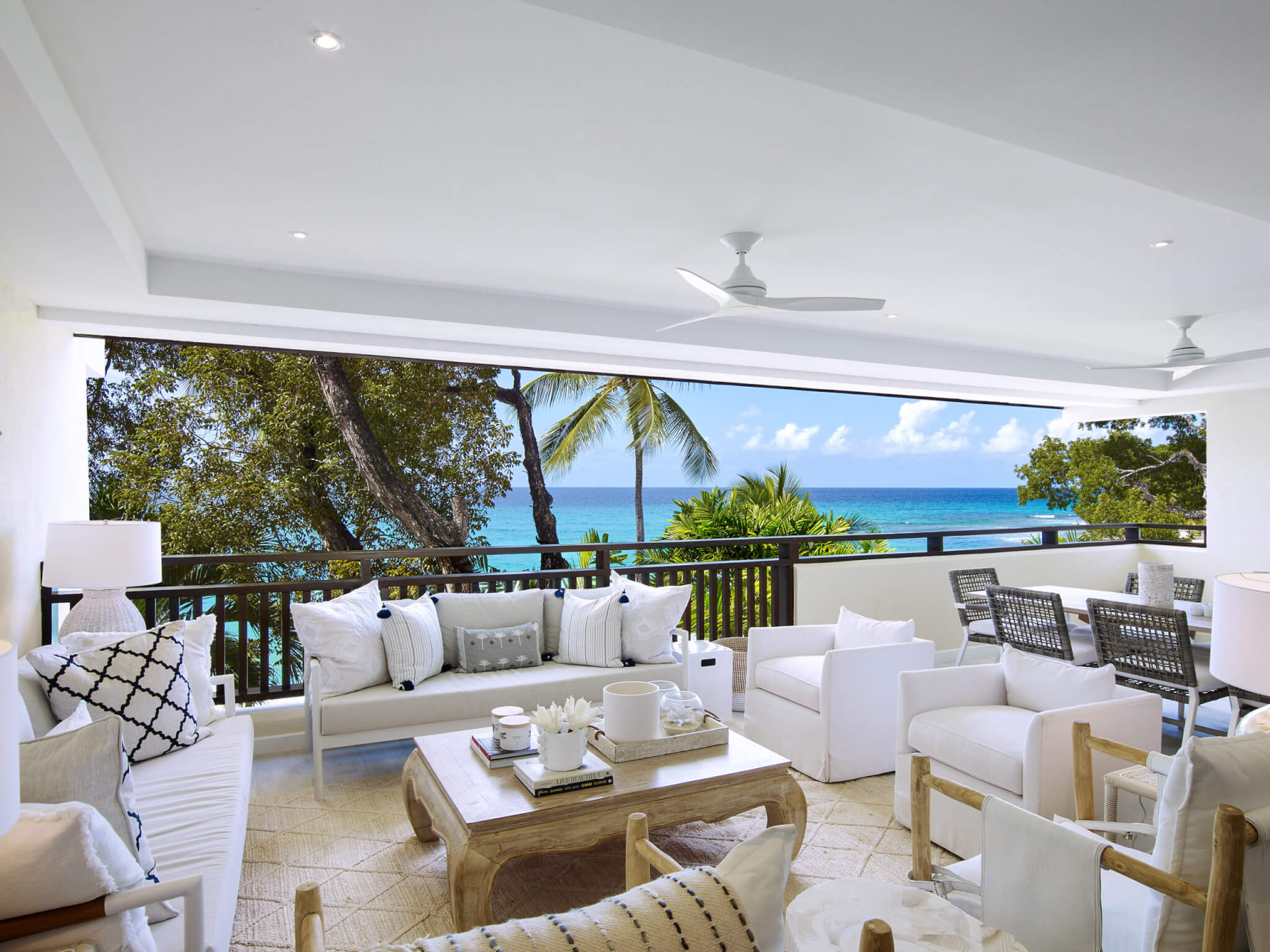 Modern, open-air living room with white sofas and wooden furniture, overlooking a balcony with a stunning view of palm trees and turquoise ocean under a sunny sky.