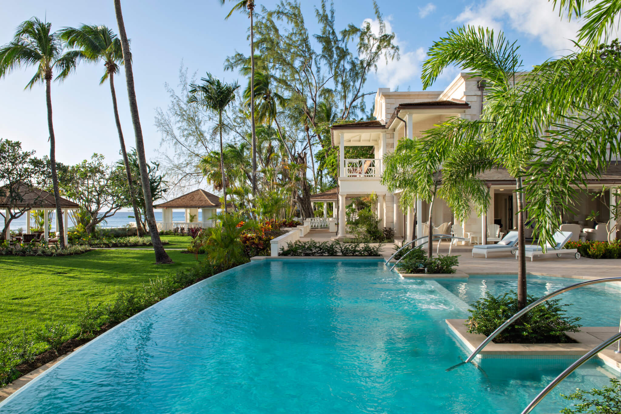 A luxurious tropical resort featuring a large outdoor swimming pool, palm trees, green lawns, sun loungers, and shaded gazebos with a view of the ocean in the background under a partly cloudy sky.