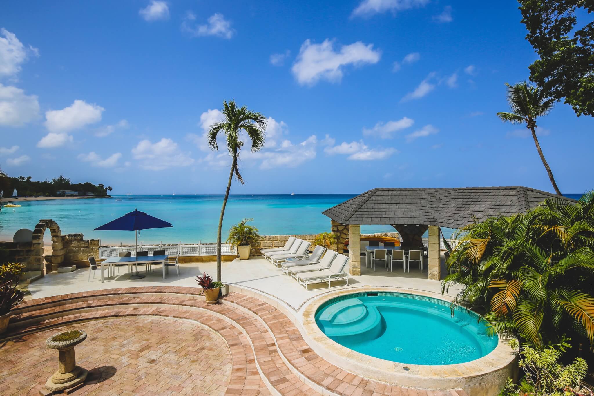 A tropical beachfront patio with a small round pool, lounge chairs, a gazebo, palm trees, and views of clear blue water under a sunny, partly cloudy sky.