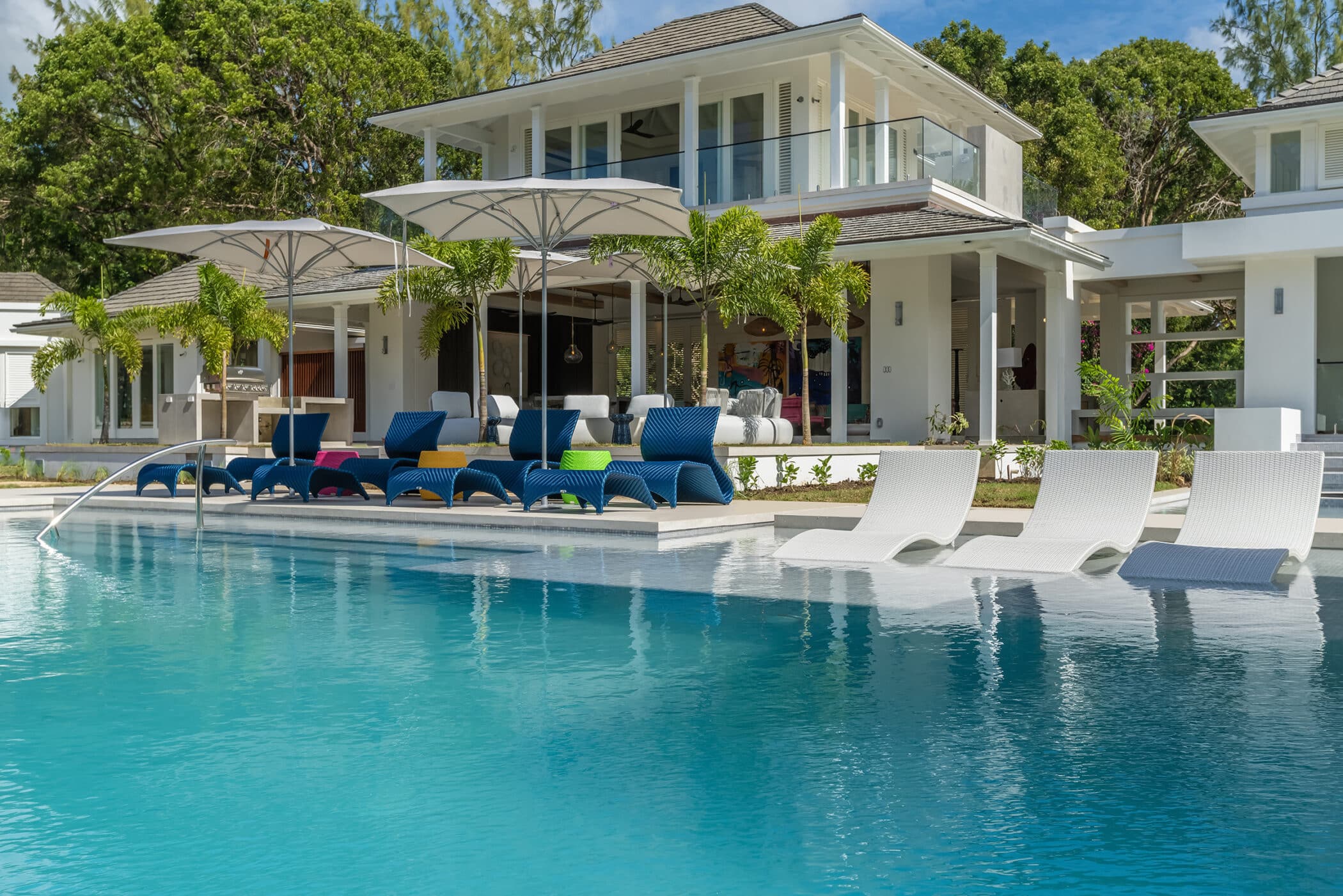 Luxurious modern house with a large glass facade, surrounded by lush green trees. In the foreground, theres a clear blue swimming pool with white and blue lounge chairs under large umbrellas on the deck.