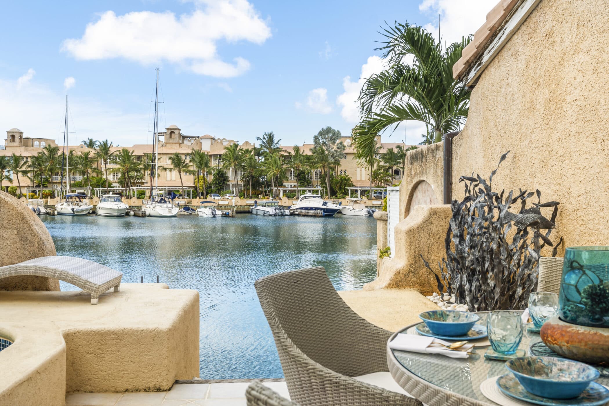 A scenic waterfront view with boats docked along a palm-lined marina. In the foreground, outdoor wicker furniture, including a table set with plates and glasses, offers a relaxing spot for dining. Stone and tropical plants add to the ambiance.