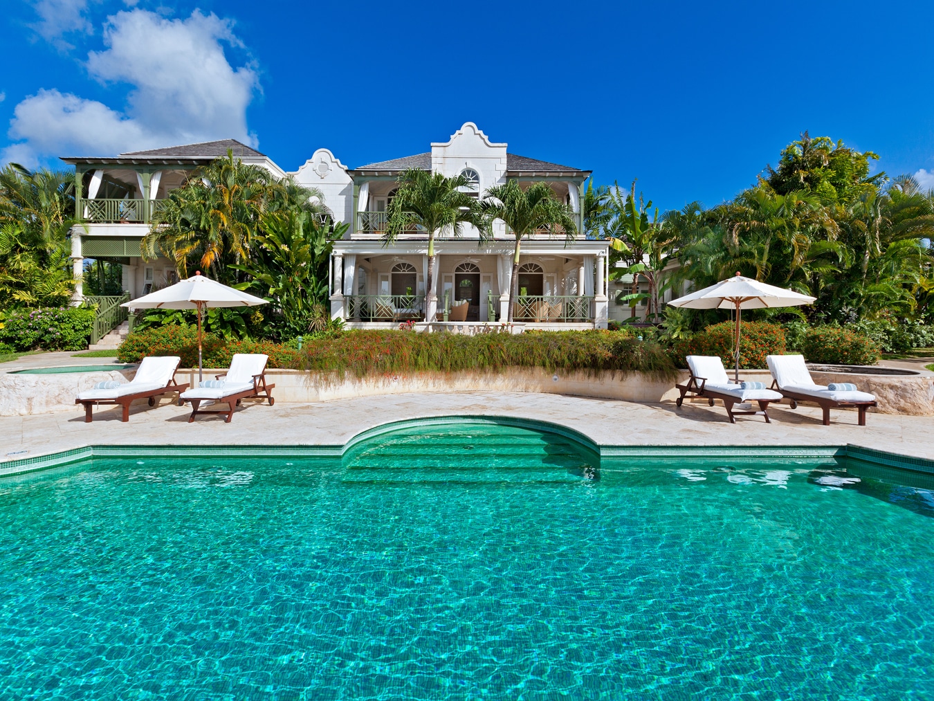 Luxurious villa with white exterior, surrounded by palm trees and lush greenery, featuring a clear turquoise swimming pool, poolside lounge chairs, and umbrellas under a bright blue sky.