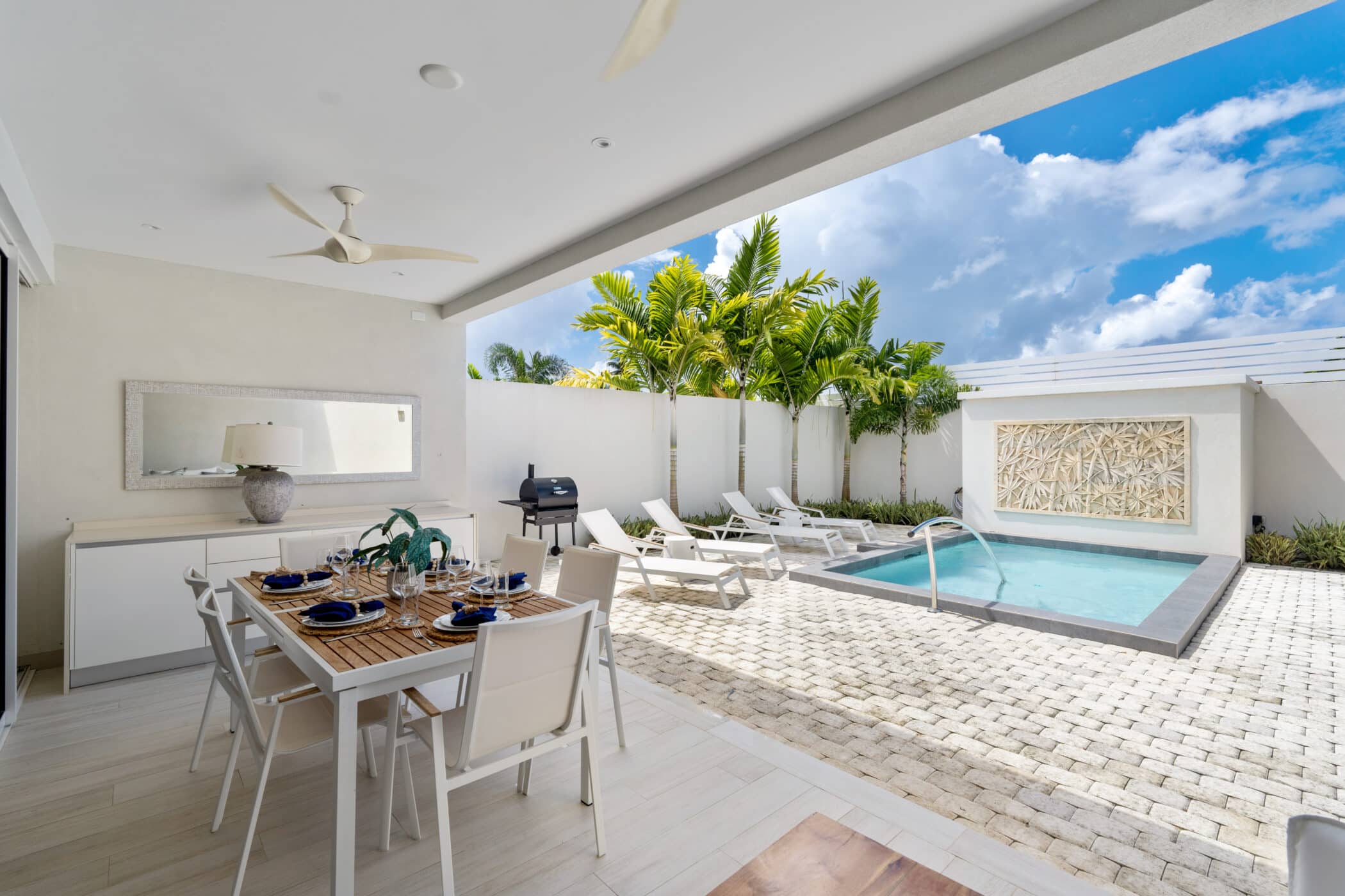 Modern outdoor patio with a dining table set for six, lounge chairs, a grill, and a small swimming pool surrounded by tropical plants under a bright blue sky with scattered clouds.