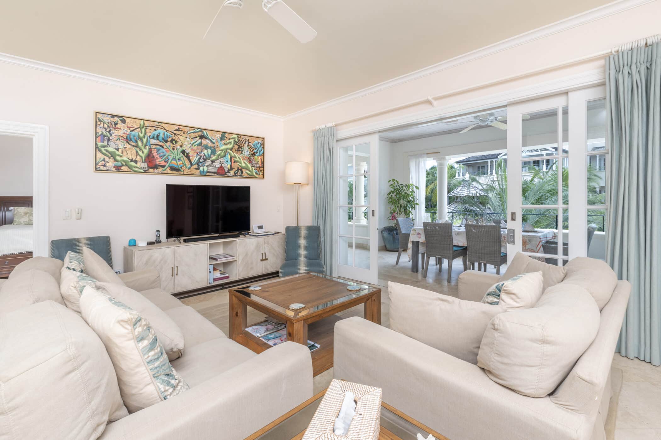 A bright, modern living room with beige sofas, a wooden coffee table, TV stand, wall art, and sliding glass doors opening to a sunlit dining area with greenery outside. Light blue curtains frame the doorway.