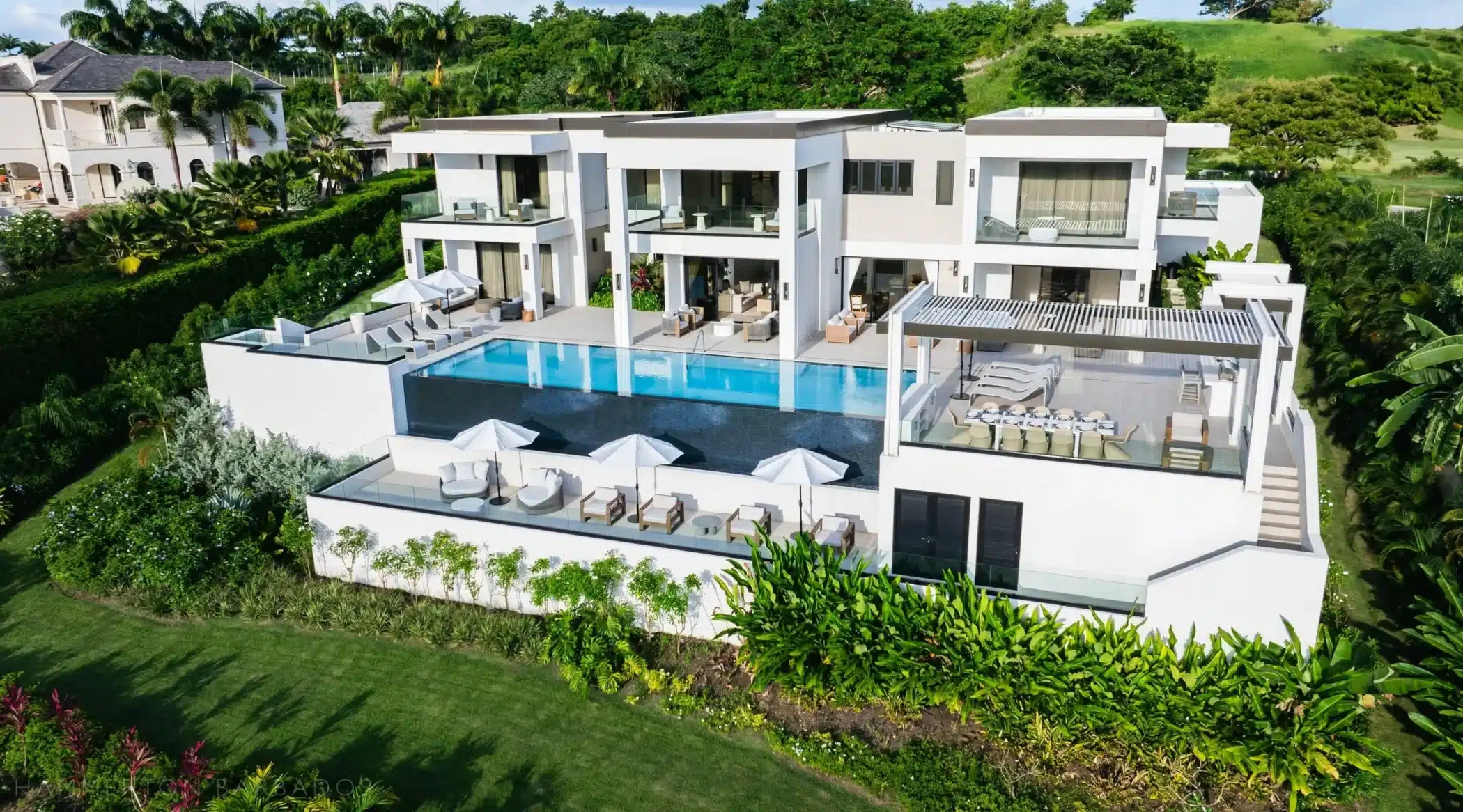 A modern luxury villa with white walls, large glass windows, and balconies, featuring an infinity pool, sun loungers, umbrellas, and lush green landscaping, set against a backdrop of tropical trees and grass.