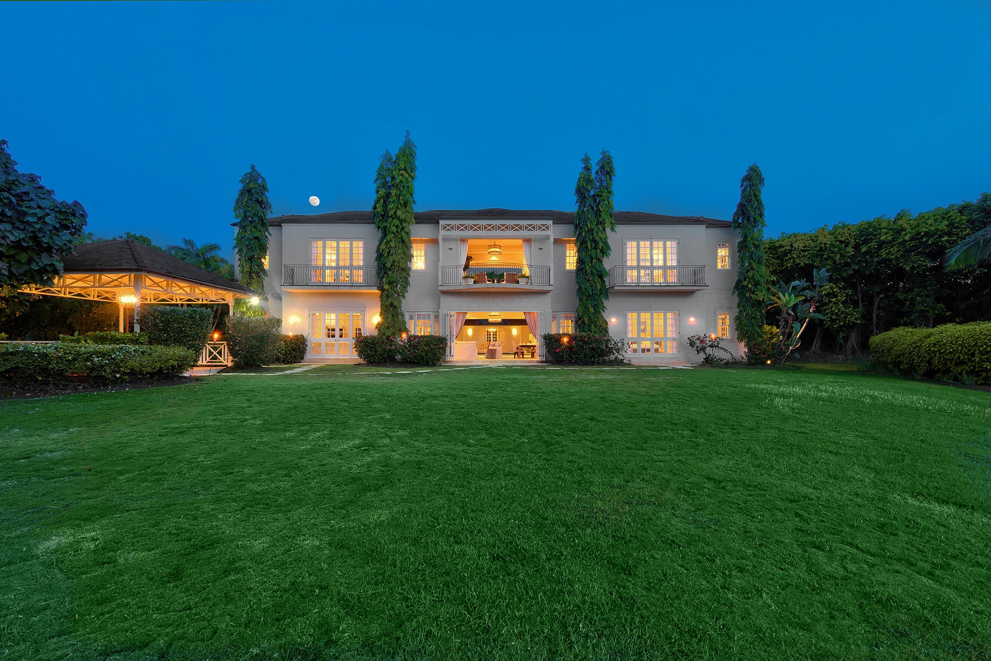 Large two-story house with tall windows and columns, warmly lit from inside at dusk, surrounded by tall trees, manicured lawn, and a gazebo on the left. The sky is clear and blue.