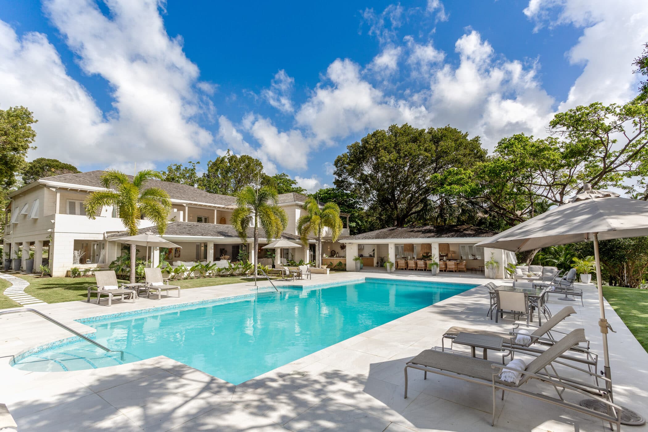 Luxury villa with a large outdoor swimming pool, sun loungers, umbrellas, and palm trees, set against a clear blue sky with scattered clouds and surrounded by lush greenery.